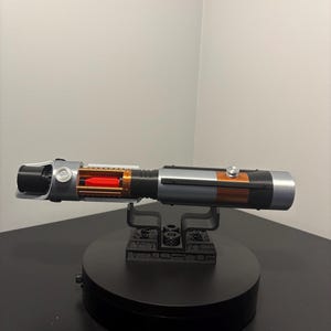 May include: A detailed replica lightsaber with a black, silver, and orange color scheme. The hilt features intricate detailing, including a visible red internal component. It rests on a black display stand, showcasing the prop's design.