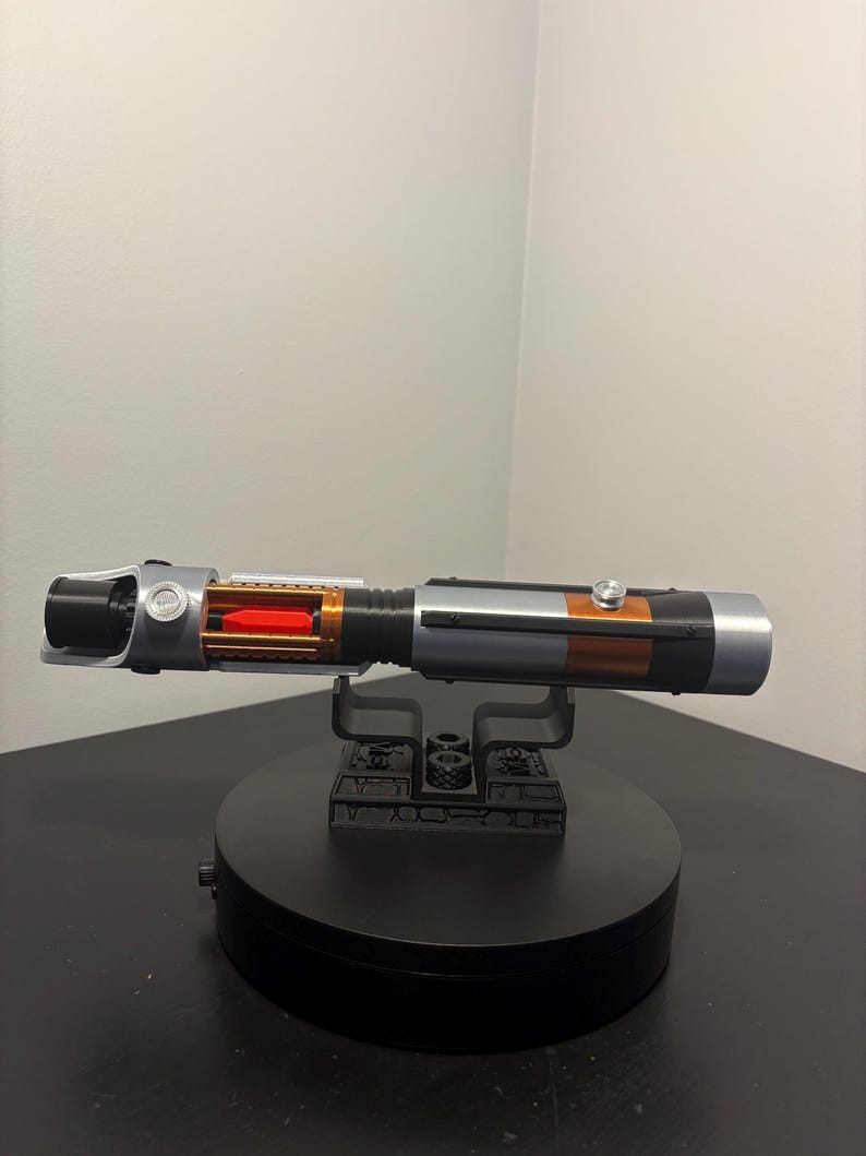 May include: A detailed replica lightsaber with a black, silver, and orange color scheme. The hilt features intricate detailing, including a visible red internal component. It rests on a black display stand, showcasing the prop's design.