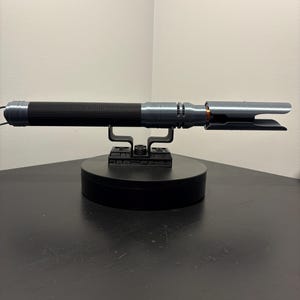 Puede incluir: Un accesorio de sable de luz plateado y negro, apoyado en un soporte de exhibici&oacute;n negro. El sable de luz presenta un mango negro acanalado y un emisor de hoja plateado. El accesorio est&aacute; dise&ntilde;ado para parecerse a un arma futurista.