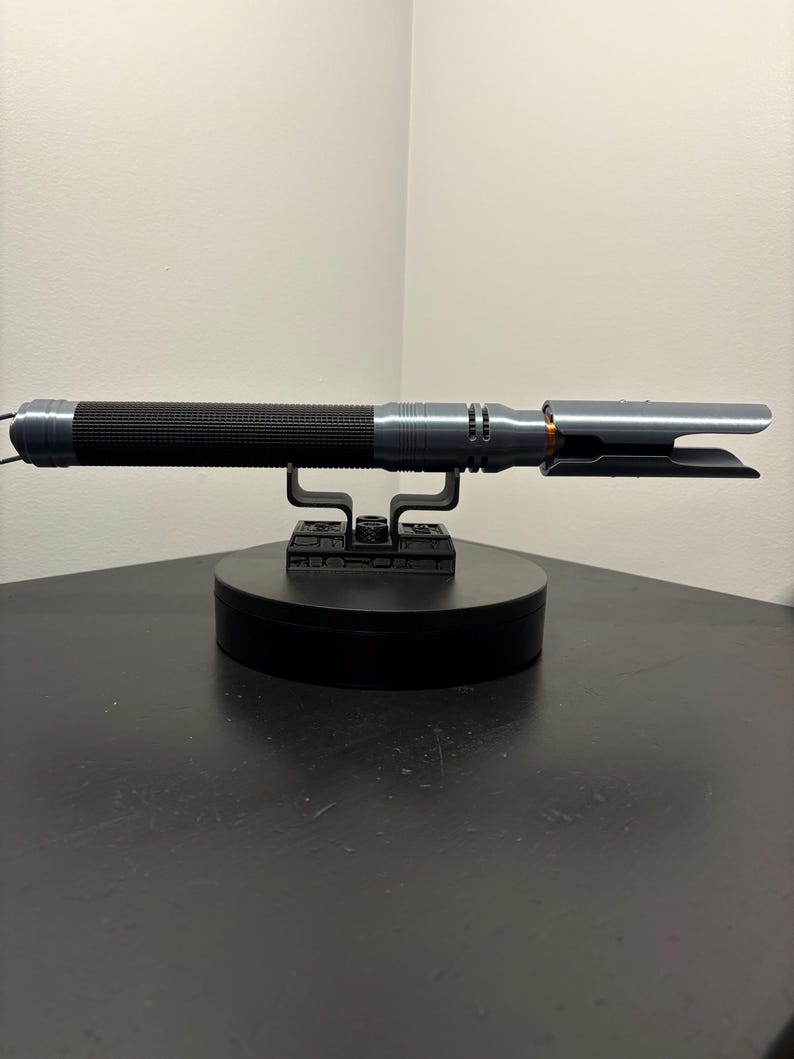 Puede incluir: Un accesorio de sable de luz plateado y negro, apoyado en un soporte de exhibici&oacute;n negro. El sable de luz presenta un mango negro acanalado y un emisor de hoja plateado. El accesorio est&aacute; dise&ntilde;ado para parecerse a un arma futurista.