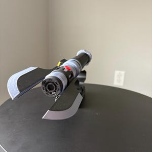 May include: A black and gray futuristic prop with a cylindrical body, red button, and silver accents. The prop has two angled blades and is set on a black surface. The design suggests a sci-fi or fantasy theme.