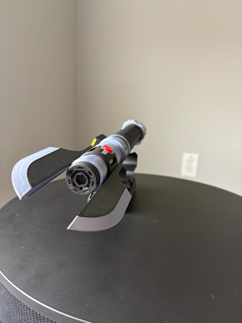 May include: A black and gray futuristic prop with a cylindrical body, red button, and silver accents. The prop has two angled blades and is set on a black surface. The design suggests a sci-fi or fantasy theme.
