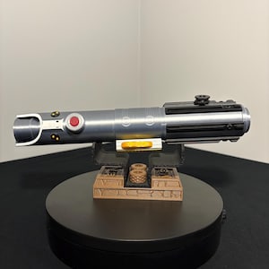 May include: A silver and black lightsaber prop with a red button and gold accents. The prop is displayed on a brown and black stand. The lightsaber is a replica of a popular science fiction weapon.