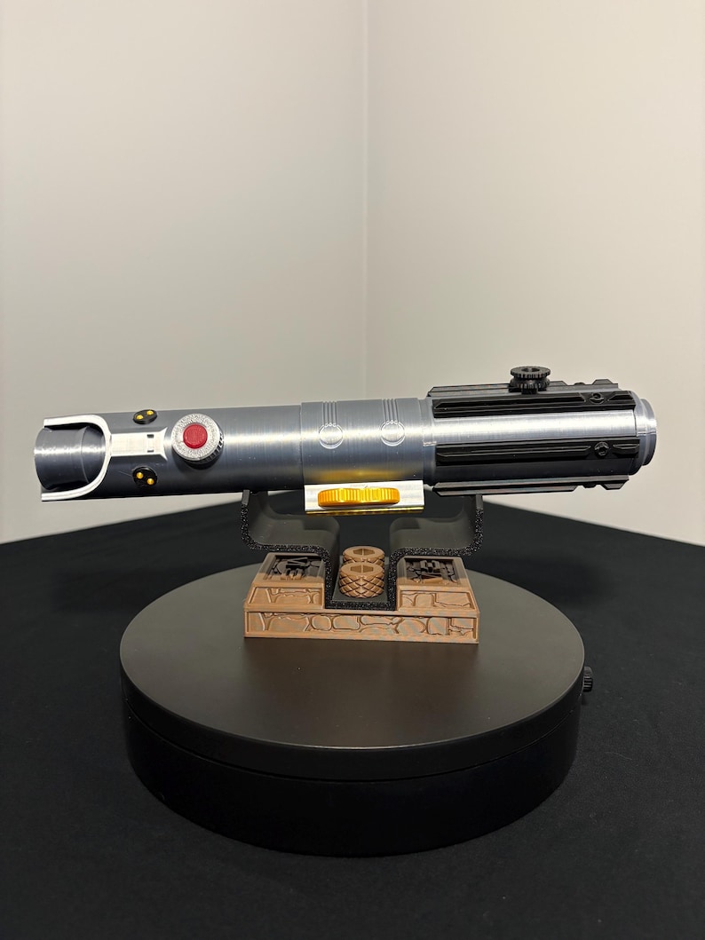 May include: A silver and black lightsaber prop with a red button and gold accents. The prop is displayed on a brown and black stand. The lightsaber is a replica of a popular science fiction weapon.