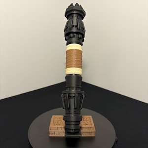 May include: A black, ornate staff with a crown-like top and a detailed base. The handle features a wrapped tan leather grip and a section of woven twine. The staff rests on a small, rectangular, brown base with carved details.