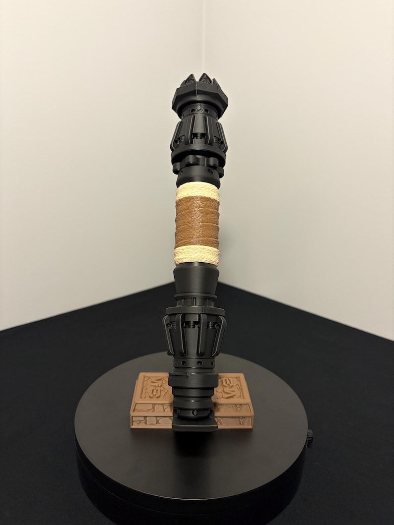 May include: A black, ornate staff with a crown-like top and a detailed base. The handle features a wrapped tan leather grip and a section of woven twine. The staff rests on a small, rectangular, brown base with carved details.