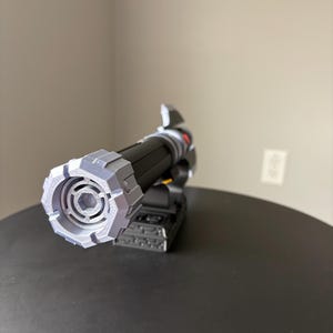 May include: A black and gray lightsaber prop with a detailed, geometric design on the hilt and emitter. The prop is resting on a black surface, showcasing its intricate construction and futuristic aesthetic. The lightsaber features a cylindrical body and a complex, angular emitter.