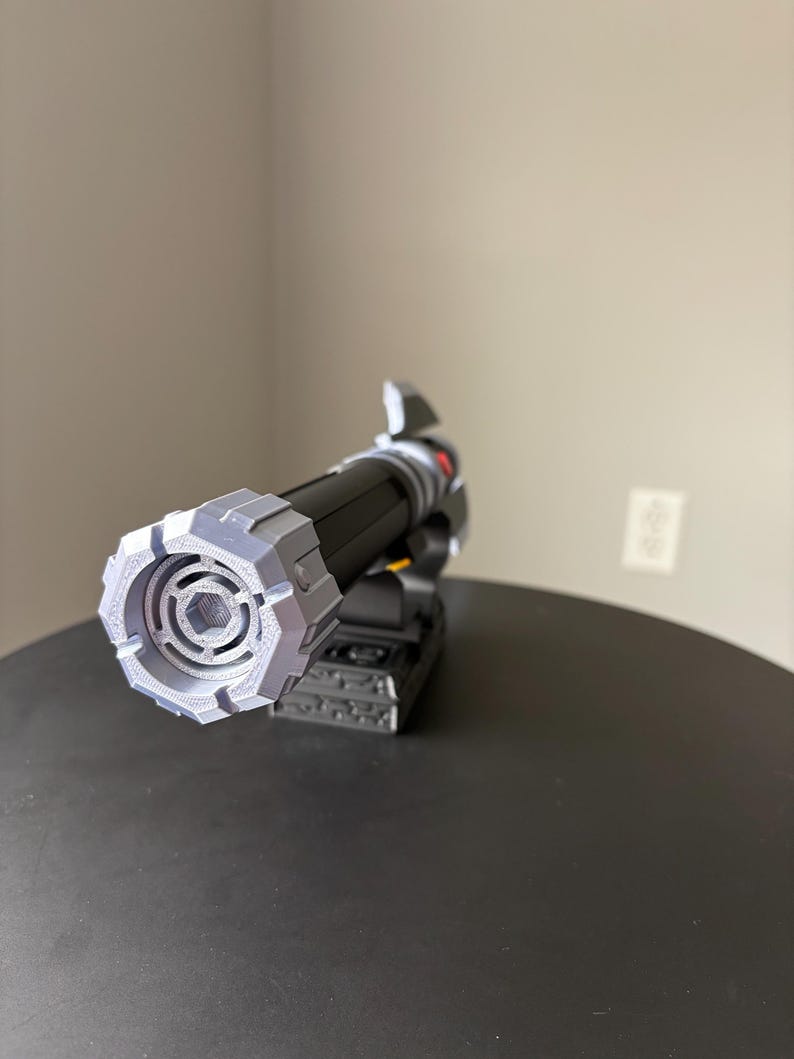 May include: A black and gray lightsaber prop with a detailed, geometric design on the hilt and emitter. The prop is resting on a black surface, showcasing its intricate construction and futuristic aesthetic. The lightsaber features a cylindrical body and a complex, angular emitter.