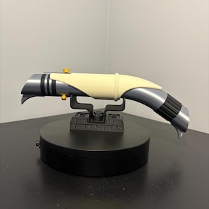 May include: A futuristic-looking, silver and black lightsaber prop with a cream-colored grip. The hilt features gold accents and is mounted on a black display stand. The lightsaber has a curved design and is likely a collectible or cosplay item.