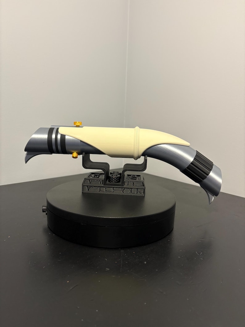 May include: A futuristic-looking, silver and black lightsaber prop with a cream-colored grip. The hilt features gold accents and is mounted on a black display stand. The lightsaber has a curved design and is likely a collectible or cosplay item.