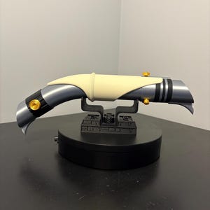May include: A futuristic lightsaber prop with a curved design. The hilt is primarily gray with black accents and gold-colored details. A cream-colored section is in the center. The lightsaber rests on a black stand, showcasing its unique shape and design.