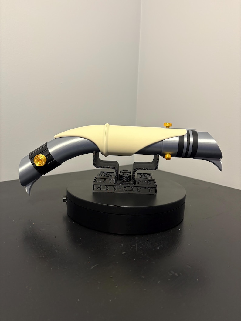 May include: A futuristic lightsaber prop with a curved design. The hilt is primarily gray with black accents and gold-colored details. A cream-colored section is in the center. The lightsaber rests on a black stand, showcasing its unique shape and design.