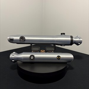 May include: Two silver-colored lightsabers are displayed on a black surface. The lightsabers have a cylindrical design with black accents and circular details. The lightsabers are resting on a black stand.