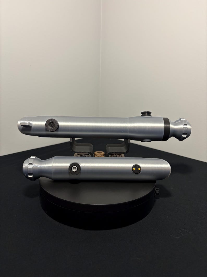 May include: Two silver-colored lightsabers are displayed on a black surface. The lightsabers have a cylindrical design with black accents and circular details. The lightsabers are resting on a black stand.