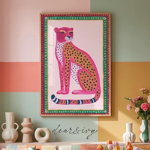 Könnte beinhalten: Ein gerahmter Wandkunstdruck mit einem stilisierten rosa Leoparden mit schwarzen Flecken, der vor einem hellrosa Hintergrund sitzt. Das Kunstwerk ist von einem dekorativen grünen und roten Rand umgeben. Der Text "dear & ivy" steht unter dem Rahmen.