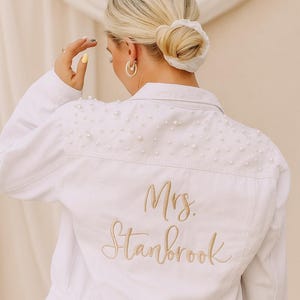 May include: White denim jacket embellished with pearls on the shoulders. The back of the jacket features the embroidered text "Mrs. Stanbrook" in a gold color. The jacket is a classic style with button closures and pockets.