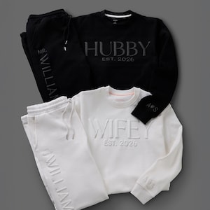 May include: Black and white matching loungewear sets. One set includes a black sweatshirt with "HUBBY EST. 2026" and matching sweatpants with "MR. WILLIAM". The other set is white with "WIFEY EST. 2026" and "WILLIAM" on the sweatpants.