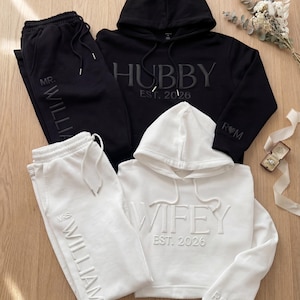 May include: Matching black and white hoodie and sweatpants set. The black hoodie says "Hubby" and the white hoodie says "Wifey". The sweatpants are embroidered with "Mr. William" and "Mrs. William".