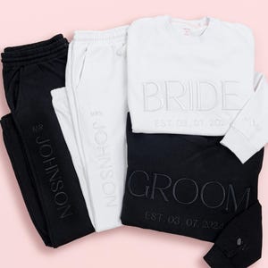 May include: A set of wedding-themed clothing in black and white. Includes sweatpants and sweatshirts, with the words "BRIDE" and "GROOM" embroidered on the tops. The date "EST. 03.07.2024" is also visible.