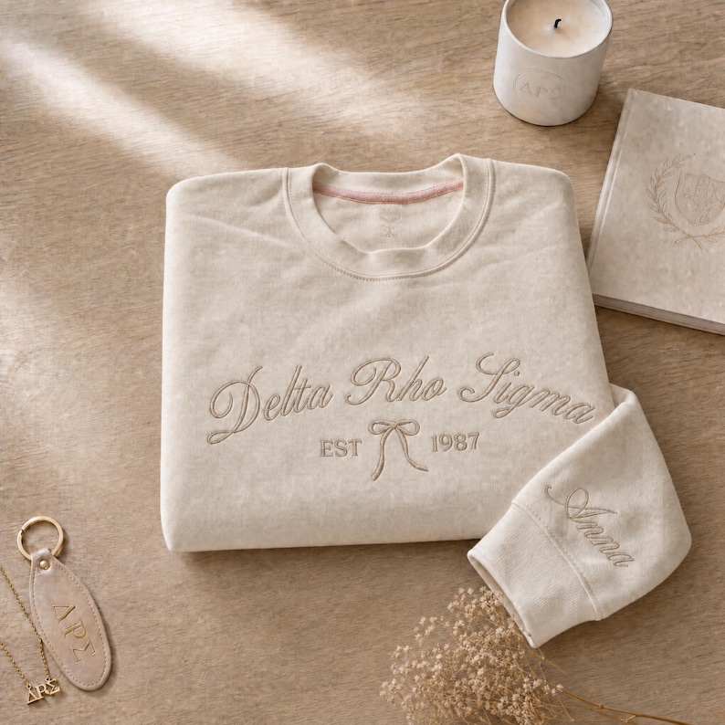 May include: A beige sweatshirt with the embroidered text "Delta Rho Sigma EST 1987" and a bow. The sleeve cuff is embroidered with the name "Anna". A candle, a book, and a keychain are also in the image.