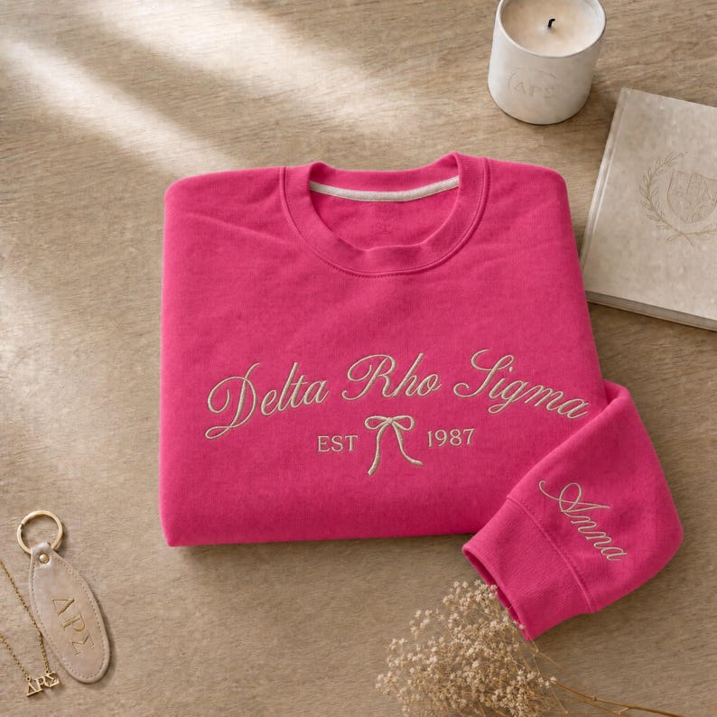 May include: A hot pink sweatshirt with the embroidered text "Delta Rho Sigma EST 1987" in a cursive font. The sleeve cuff is embroidered with the name "Anna". A gold-colored keychain and a small candle are also visible.