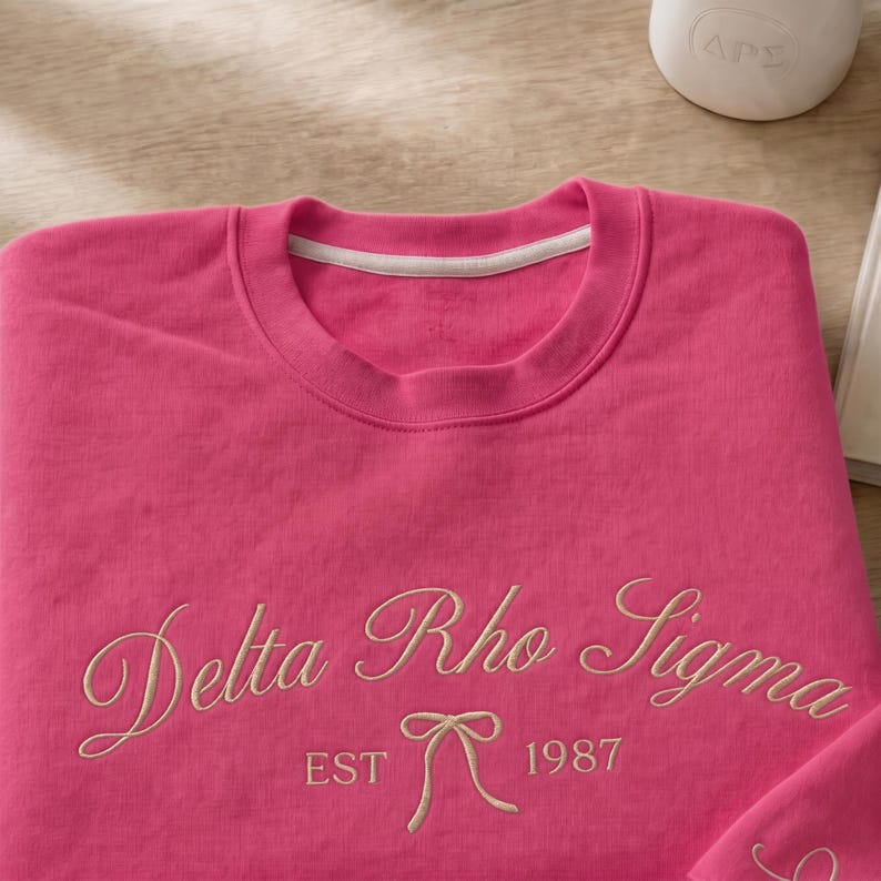 May include: A bright pink sweatshirt with the embroidered text "Delta Rho Sigma EST 1987" in a cursive font. The sweatshirt has a crew neck and a white trim. The design includes a bow.