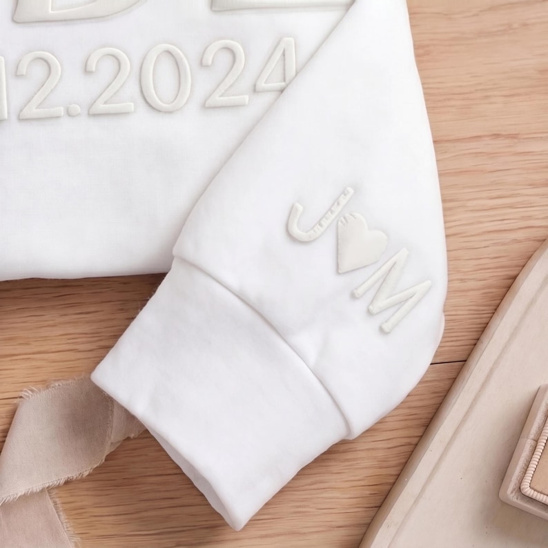 May include: White sweatshirt with raised lettering and the date "12.2024". The sleeve cuff displays the initials "J♡M" in a similar raised design. The garment is made of a soft, likely cotton material.