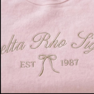 May include: A light pink sweatshirt with embroidered text in a cursive font. The text reads "Delta Rho Sig" with "EST" and "1987" below a bow graphic. The embroidery is in a light tan color.