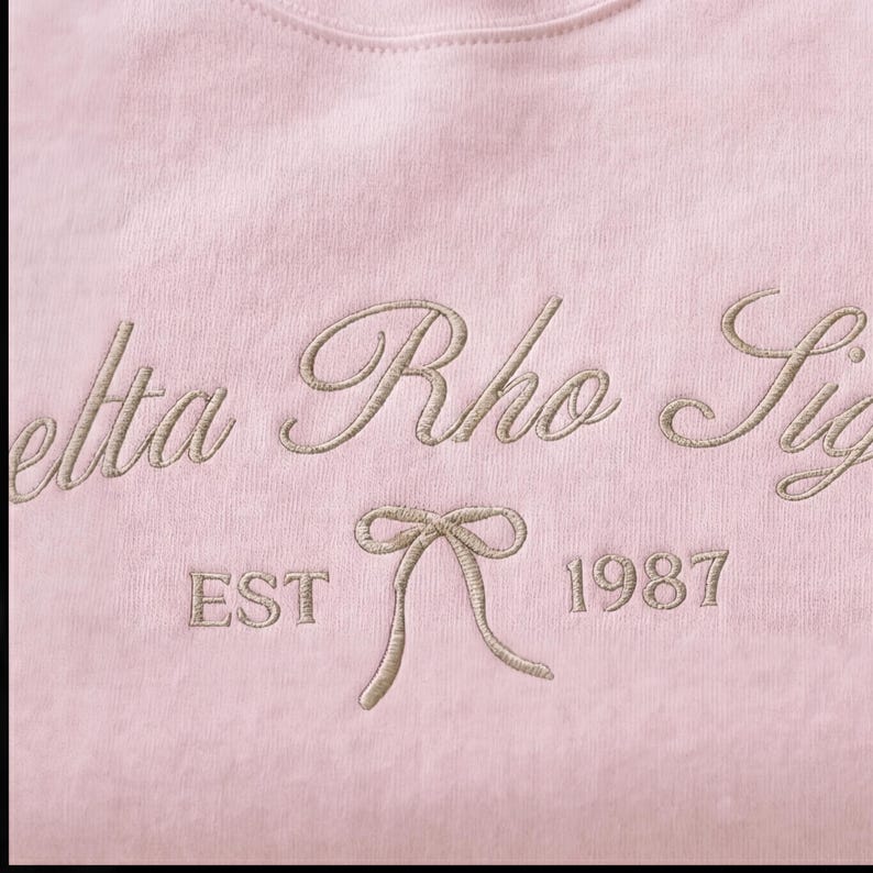 May include: A light pink sweatshirt with embroidered text in a cursive font. The text reads "Delta Rho Sig" with "EST" and "1987" below a bow graphic. The embroidery is in a light tan color.