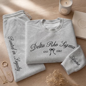 May include: A light gray sweatshirt and sweatpants set with black embroidered text. The sweatshirt features "Delta Rho Sigma EST 1987" and a bow. The sweatpants say "Rooted in Loyalty". A candle, a book, and a keychain are also present.