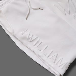 May include: White sweatpants with a drawstring and the embossed name "WILLIAM". A white sweatshirt with the embossed name "WILLIAM" is also visible. The clothing is made of a soft material.
