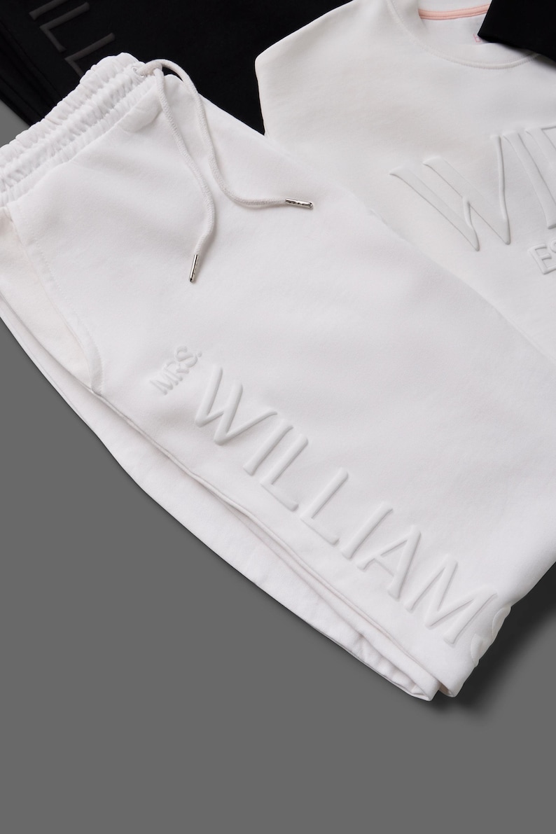 May include: White sweatpants with a drawstring and the embossed name "WILLIAM". A white sweatshirt with the embossed name "WILLIAM" is also visible. The clothing is made of a soft material.