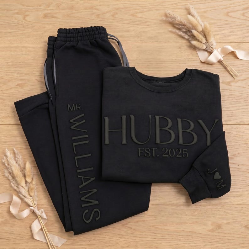 May include: Black matching loungewear set featuring a sweatshirt and sweatpants. The sweatshirt has the word "HUBBY" and "EST. 2025" printed on it. The sweatpants have "MR. WILLIAMS" printed on the side. A small heart and initials are on the sleeve.