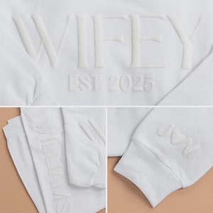 May include: White sweatshirt with the word "WIFEY" and "EST. 2025" embossed on the front. The sleeve features initials "J♡M". Matching white pants with "MRS WILLIAMS" embossed on the side. A bridal-themed apparel set.