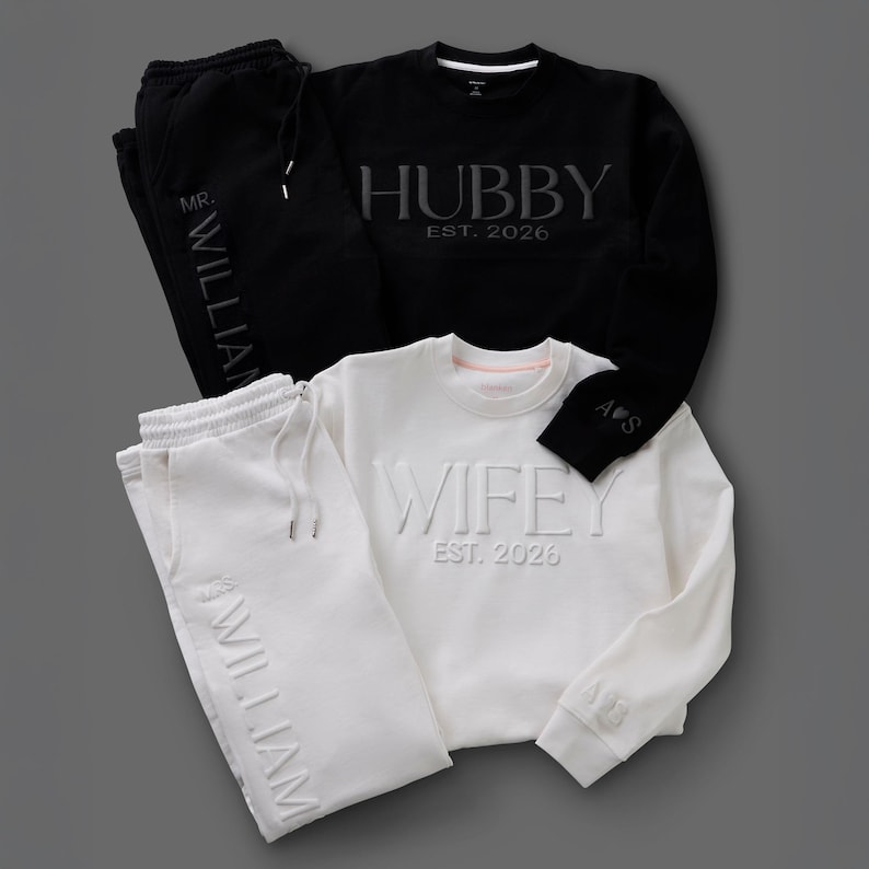 May include: Black and white matching loungewear sets. One set includes a black sweatshirt with "HUBBY EST. 2026" and matching sweatpants with "MR. WILLIAM". The other set is white with "WIFEY EST. 2026" and "WILLIAM" on the sweatpants.