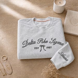 May include: A light gray sweatshirt with the embroidered text "Delta Rho Sigma EST 1987" in black script. The sleeve cuff is embroidered with the name "Anna". A candle, book, and keychain are also visible.