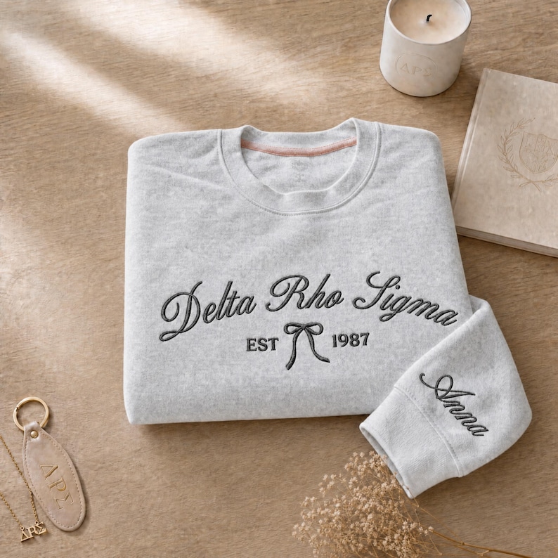 May include: A light gray sweatshirt with the embroidered text "Delta Rho Sigma EST 1987" in black script. The sleeve cuff is embroidered with the name "Anna". A candle, book, and keychain are also visible.