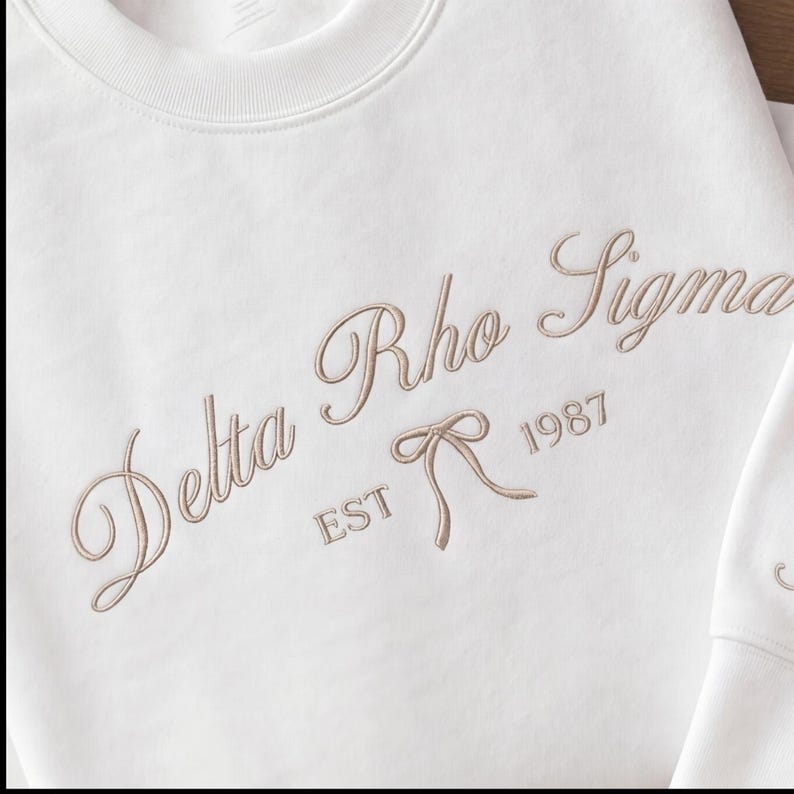May include: A white sweatshirt with embroidered text in a cursive font. The text reads "Delta Rho Sigma" with "EST 1987" below a bow graphic. The sweatshirt is a crew neck style.