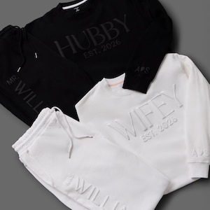 May include: Black and white matching loungewear sets. The black set includes a sweatshirt with "HUBBY EST. 2026" and matching sweatpants. The white set features a sweatshirt with "WIFEY EST. 2026" and matching sweatpants. Both sets have the initials "A&S".