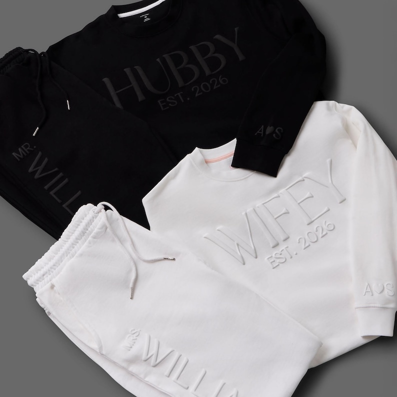 May include: Black and white matching loungewear sets. The black set includes a sweatshirt with "HUBBY EST. 2026" and matching sweatpants. The white set features a sweatshirt with "WIFEY EST. 2026" and matching sweatpants. Both sets have the initials "A&S".