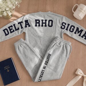 May include: A gray sweatshirt and sweatpants set with the Greek letters "DELTA RHO SIGMA" in navy blue. The sweatpants have the text "ROOTED IN LOYALTY" printed vertically on the leg. A white mug and a navy blue book are also visible.