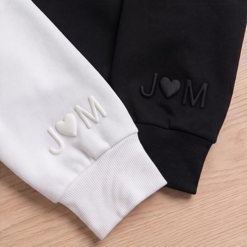 May include: Close-up of two sweatshirts, one white and one black. Both sleeves feature the initials "J♡M" in a raised, textured design. The white sweatshirt has a white heart, while the black sweatshirt has a black heart.