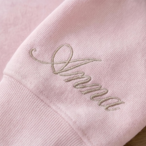 May include: Close-up of a light pink sweatshirt sleeve with the embroidered name "Anna" in a cursive font. The thread color is a muted, neutral tone, creating a subtle contrast against the soft fabric.