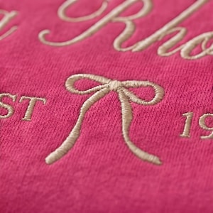 May include: Close-up of a hot pink fabric with embroidered text and a bow design. The text appears to be a name and the year 19. The embroidery is a light beige color, creating a contrast against the pink background.