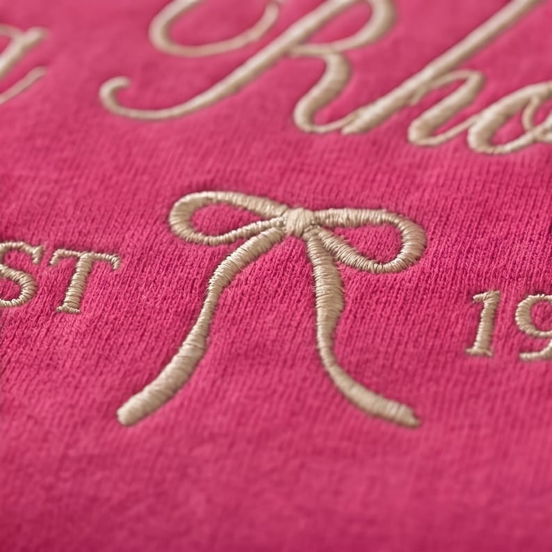 May include: Close-up of a hot pink fabric with embroidered text and a bow design. The text appears to be a name and the year 19. The embroidery is a light beige color, creating a contrast against the pink background.