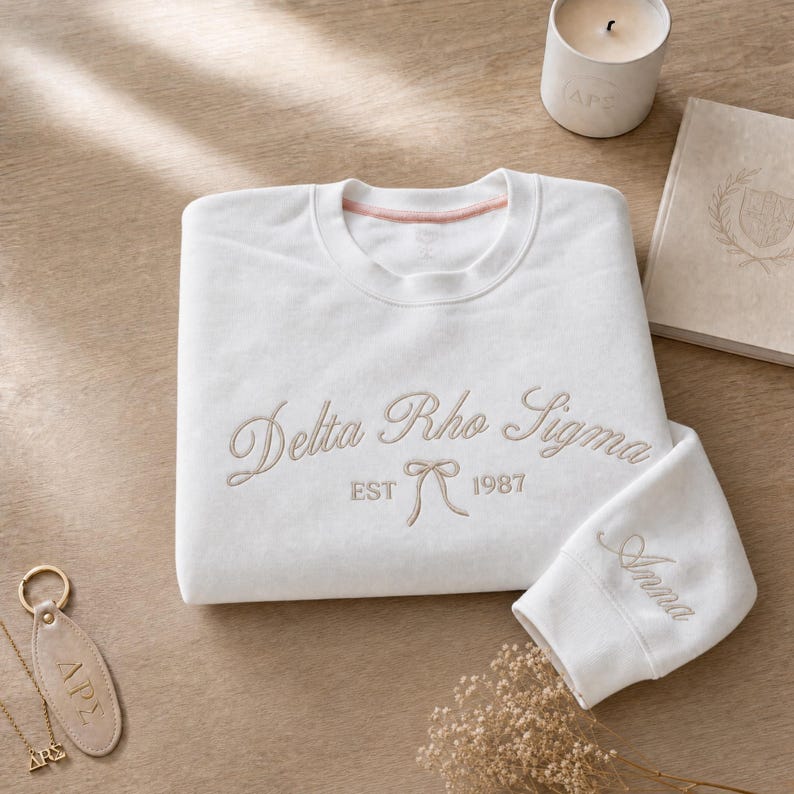 May include: A white sweatshirt with the embroidered text "Delta Rho Sigma EST 1987" in a cursive font. The sleeve has the name "Anna" embroidered. A leather keychain and necklace with "&Delta;&Rho;&Sigma;" are also visible.