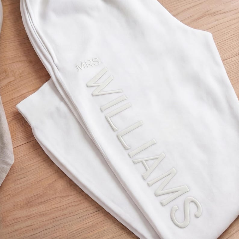 May include: White sweatpants with the name "WILLIAMS" and the title "MRS" in a raised, tonal font. The pants are folded on a light wood surface.