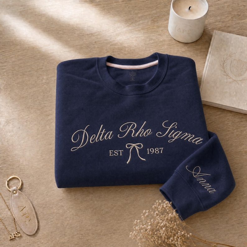 May include: Navy blue sweatshirt with the embroidered text "Delta Rho Sigma EST 1987" in a cursive font. The sleeve cuff has the name "Anna" embroidered. A gold-colored keychain and necklace are visible.