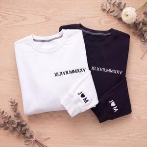 May include: A white sweatshirt and a black sweatshirt, each featuring the Roman numeral date XI.XVII.MMXXV printed on the chest and the letters J and M separated by a heart symbol embroidered on the sleeve cuff, showcasing a matching set.