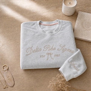 May include: A light gray sweatshirt with the embroidered text "Delta Rho Sigma" and "EST 1987" in a cursive font. The sleeve cuff is embroidered with the name "Anna". A gold keychain and necklace are visible.